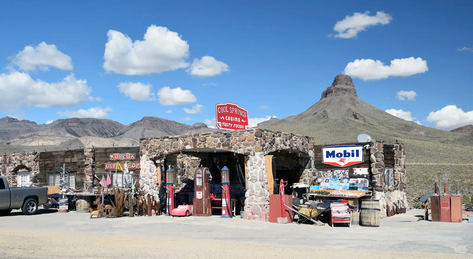 Cobblestone structure with gas pumps in front on the side of a dirt road with a sign that reads 'Cool Springs-Cabins-Tasty Foods'.