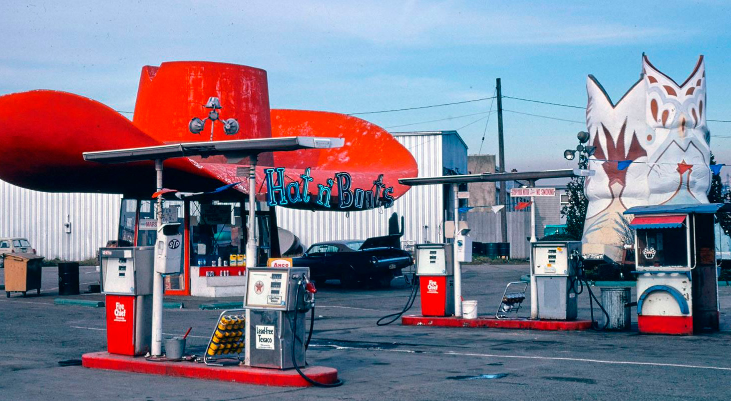 Giant red cowboy hat and white cowboy boots statues in a gas station parking lot that reads 'Hat n' Boots'.