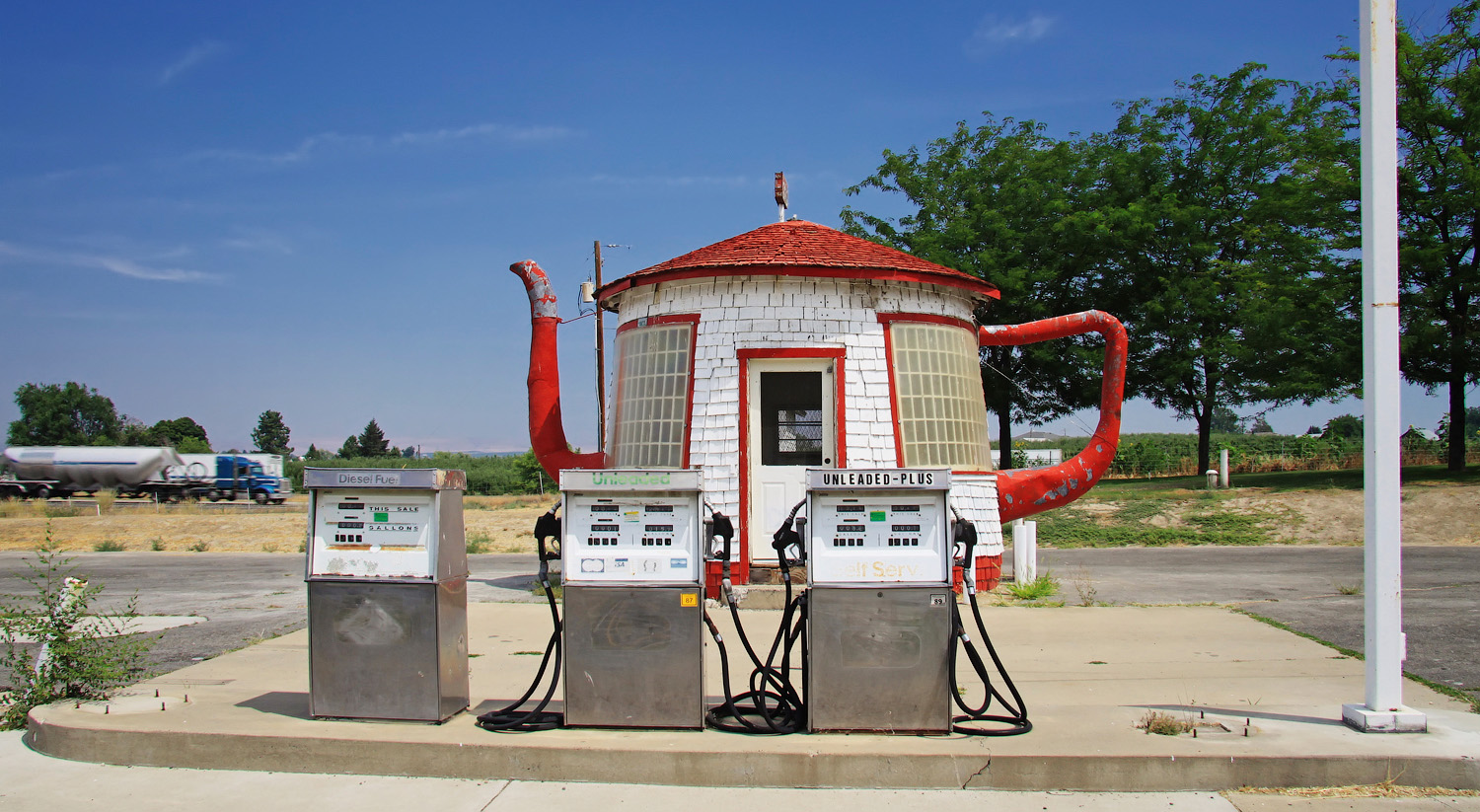 Red and white teapot shaped building in the background of three old silver gas pumps.