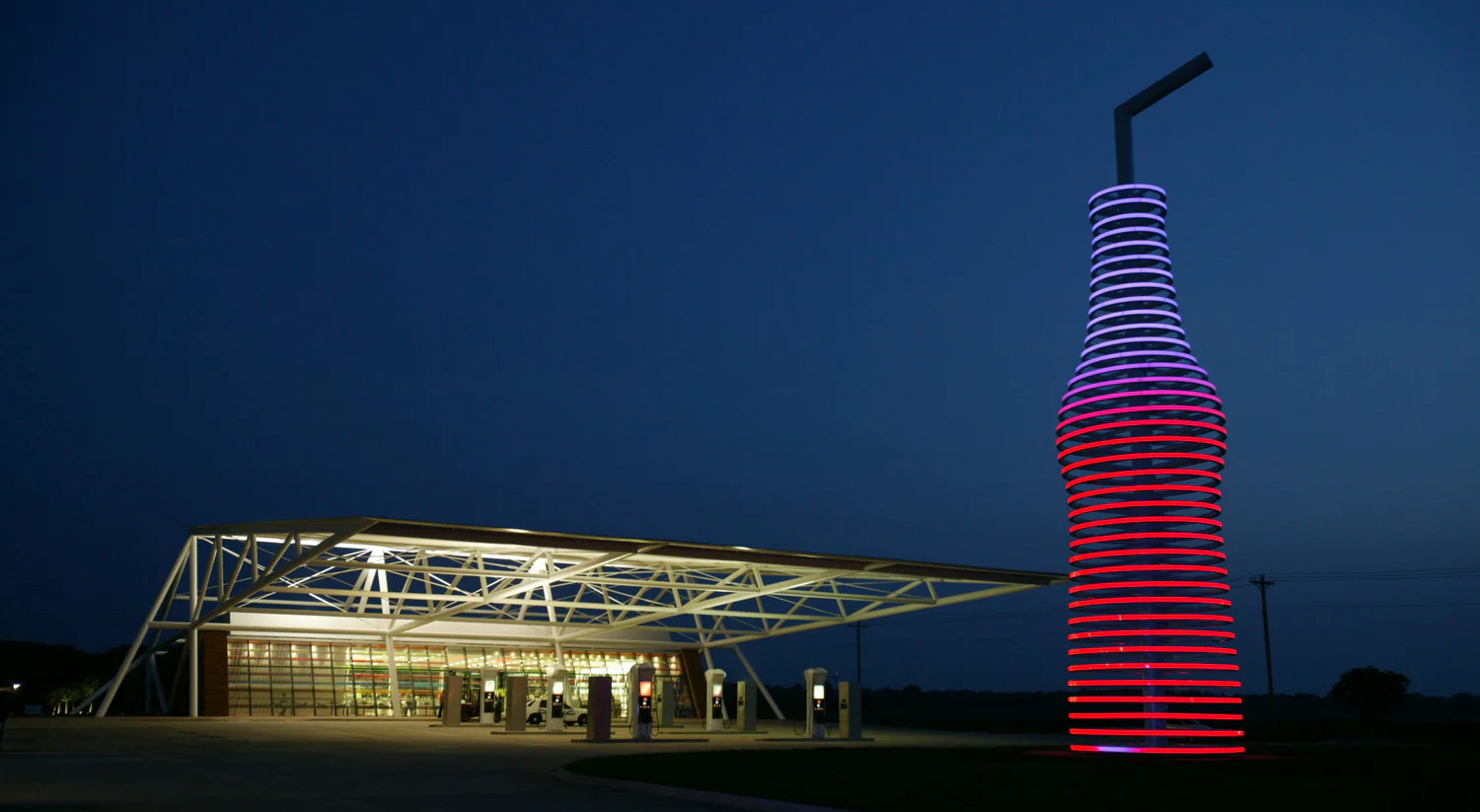 Isolated gas station building with many windows, a geometrical, open roof frame, and a large, minimalist, glowing sculpture of a soda bottle.